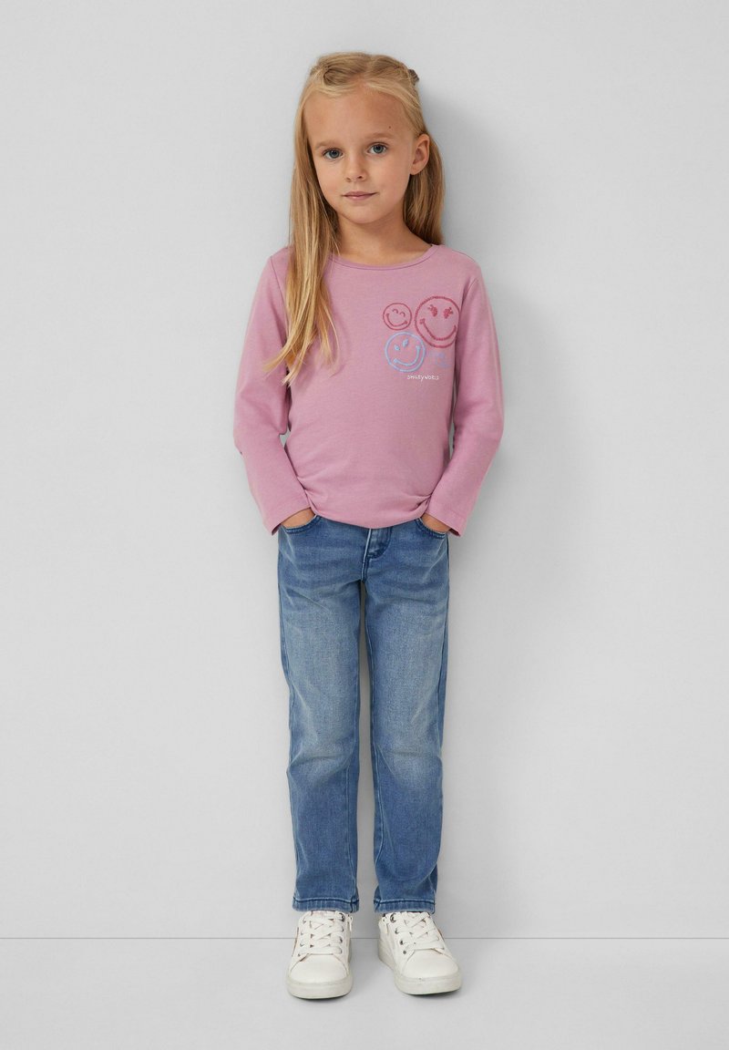 Junges Mädchen mit langen blonden Haaren, das ein pinkes Langarmshirt mit Smiley-Gesichtern, blaue Jeans und weiße Sneakers trägt, steht mit den Händen in den Taschen.