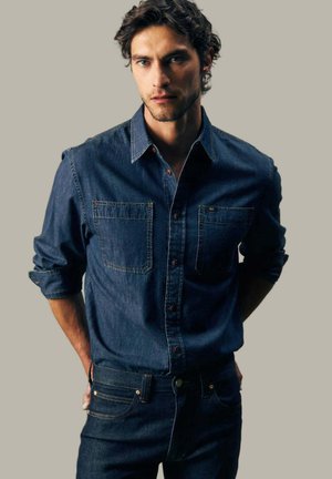 Junger Mann mit dunkel lockigem Haar, trägt ein hochgekrempeltes langärmeliges Jeanshemd und dunkle Jeans, steht vor schlichtem Hintergrund.