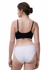 Woman with brown hair tied in a bun wearing a black bra and white high-waisted panties with lace trim, shown from the back against white background.