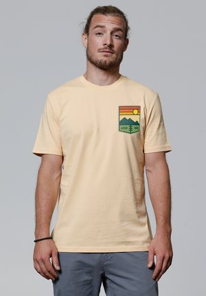 Korte mouwen, lichtgele katoenen t-shirt met een grafisch pocketontwerp featuring bergen, een zon en de tekst "GOOD VIBE."
