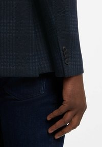 Ciemnoskóra dłoń spoczywająca obok rękawa granatowej marynarki w kratę z czterema czarnymi guzikami, nad kieszenią ciemnoniebieskich dżinsów.