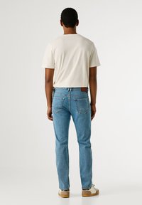 Jasnobeżowa koszulka z krótkim rękawem o okrągłym dekolcie, zestawiona z dopasowanymi niebieskimi dżinsami, które mają klasyczny pięciokieszeniowy krój i skórzaną naszywkę.