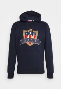 Sudadera azul marino que presenta un gran escudo bordado con acentos en rojo, blanco y dorado, un bolsillo frontal y una capucha con cordón.