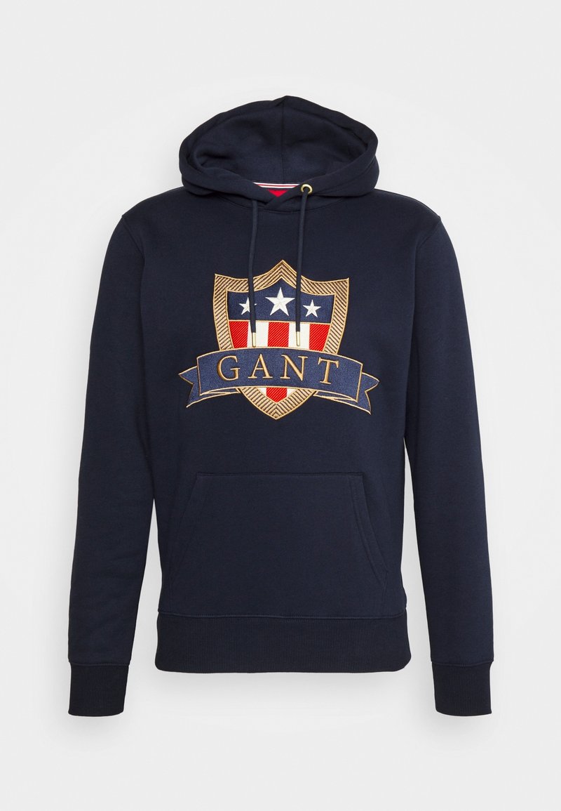 Sudadera azul marino que presenta un gran escudo bordado con acentos en rojo, blanco y dorado, un bolsillo frontal y una capucha con cordón.