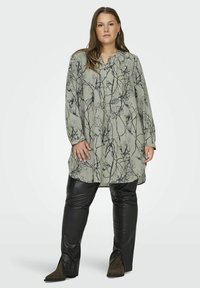 Tunique vert clair avec imprimé botanique noir, manches longues et encolure en V ; associée à un pantalon noir en simili cuir et des bottines brunes.