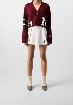 Kvinde iført en kort, bordeauxfarvet cardigan med hvide striber og nummerlapper, en hvid plisseret mininederdel, beige sokker og spidse sko.