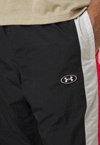 Black athletic pants with a shiny texture, featuring a white side stripe and an Under Armour logo. The fabric appears lightweight and smooth.