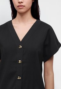 Femme portant un chemisier noir à col en V, manches courtes et trois boutons dorés distinctifs sur le devant.
