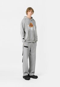Sudadera con capucha gris, con gráficos frontales y pantalones a juego. Presenta un tejido texturizado, bolsillos laterales y detalles en negro. Calzado negro.