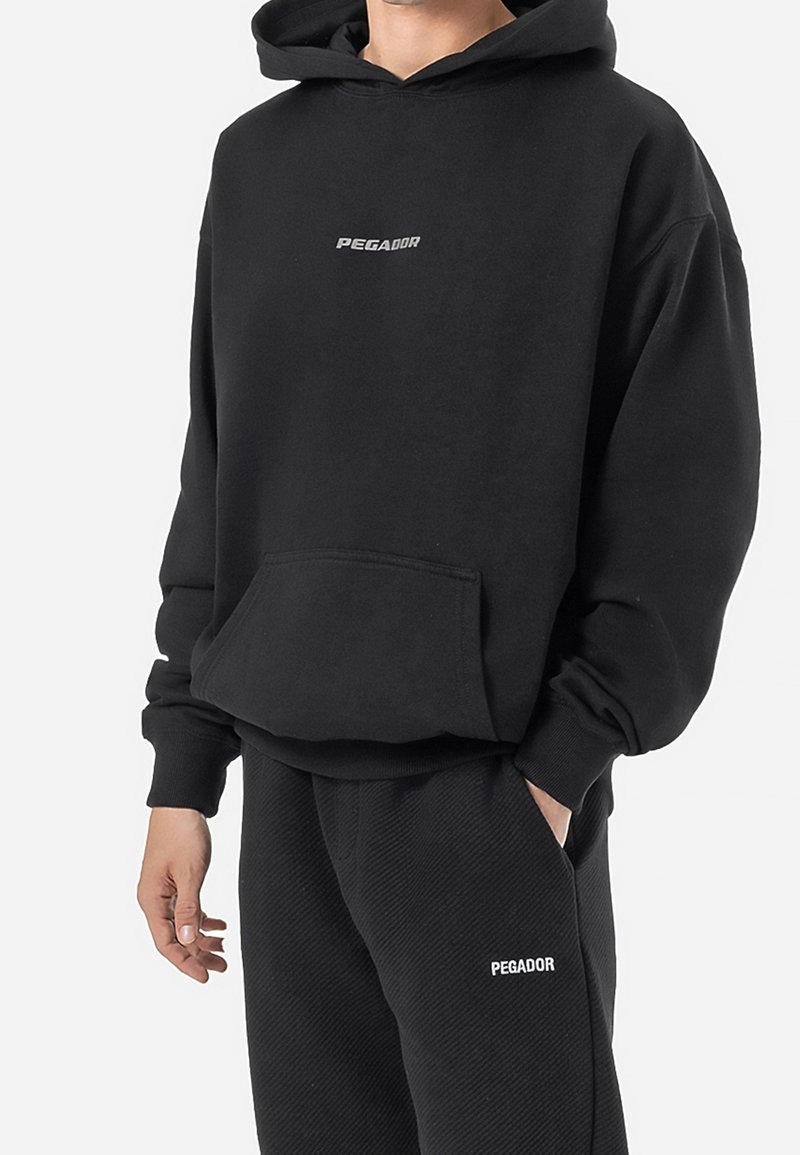 Personne portant un sweat à capuche noir et un pantalon noir, tous deux avec le logo "PEGADOR", debout avec une main dans la poche et l'autre détendue le long du corps.