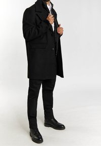 Abrigo de lana negro con un gran cuello y dos bolsillos delanteros, superpuesto a una chaqueta de plumas negra con cremallera. Pantalones negros y botas completan el conjunto.