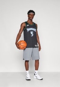 Camisola de basquetebol preta com o gráfico "TIMBERWOLVES" e o número 5. Calções cinzentos, meias brancas e sapatilhas da Nike. Segurando uma bola de basquetebol laranja.