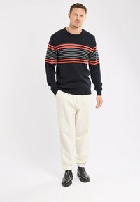 Homme portant un pull noir avec des rayures horizontales rouges et blanches, un pantalon crème et des bottes noires, se tenant avec une main dans la poche.