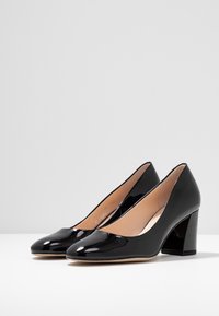 Czarne lakierowane szpilki na wysokim obcasie z zaokrąglonym noskiem i pełnym obcasem. Gładka tekstura z naturalną wkładką. Klasyczny design z minimalistycznymi akcentami.