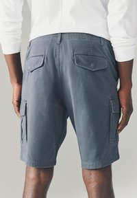 Shorts cargo gri cu buzunare laterale, cu o croială relaxată și o textură netedă, tăiați deasupra genunchiului. Buzunarele de la spate cu clapete sunt vizibile.