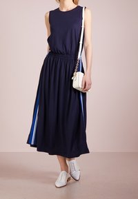 Donna che indossa un abito midi senza maniche di colore blu navy con strisce laterali blu e bianche, borsa a tracolla bianca e scarpe bianche con lacci.