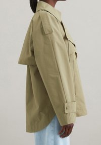 Olivengrøn bomuldsjakke med en boxy pasform, skulderepauleter, store forlommer og en buet nederkant. Har knaplukning ved manchetterne og et minimalistisk design.