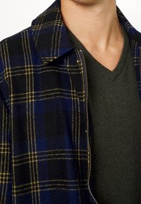 Mann trägt einen dunkelgrünen V-Ausschnitt-Pullover unter einer blau-schwarz karierten Jacke mit gelben Streifen und einem Reißverschluss.