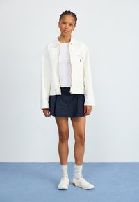 Weißer Reißverschlussjacke mit Tasche, getragen über einem weißen Oberteil. Marineblaue Skirt mit fransigem Saum, weiße Sneakers und gerippte Socken runden den Look ab.