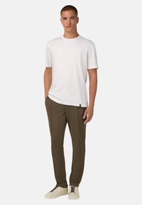 Balts īsām piedurknēm T-krekls apvienots ar olīvzaļiem slim-fit biksēm un bēžām sporta kurpēm ar tumši brūnām šņorēm. Modelis stāv pret vienkāršu fonu.