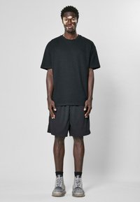 T-shirt en coton noir et shorts en maille noirs, tous deux oversize. Le modèle porte des baskets grises avec des lacets blancs et un accent texturé.