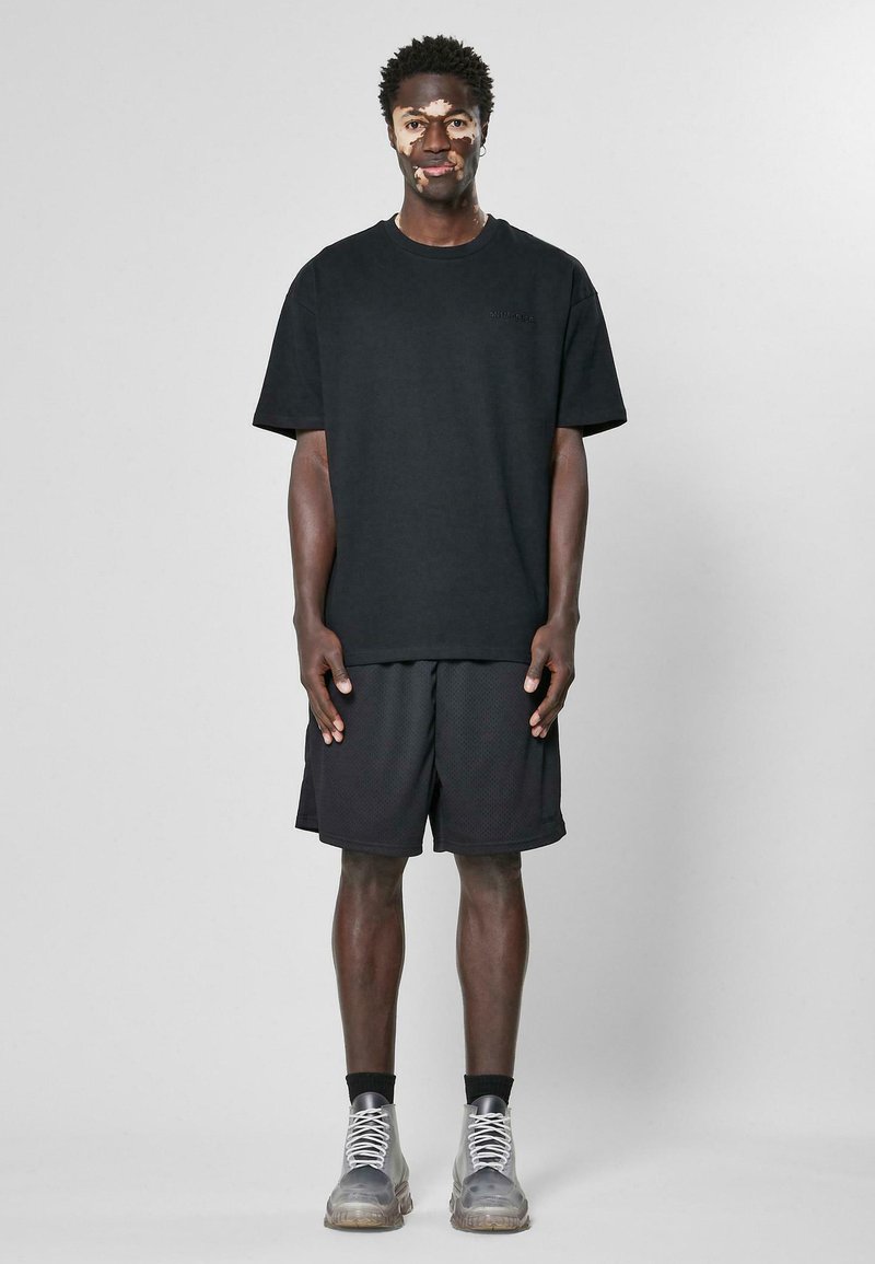 T-shirt en coton noir et shorts en maille noirs, tous deux oversize. Le modèle porte des baskets grises avec des lacets blancs et un accent texturé.