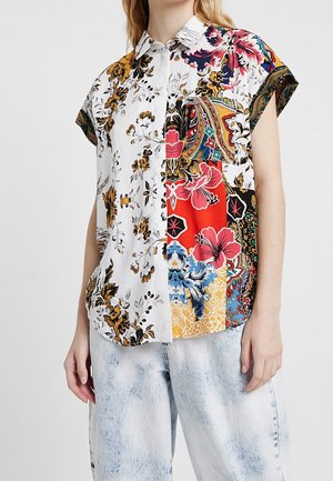 Femme portant une chemise à manches courtes boutonnée avec des motifs floraux et cachemire mélangés en blanc, rouge, bleu et jaune, associée à un jean délavé clair.