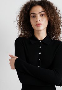 Haut à manches longues noir côtelé avec un col, présentant une patte de boutonnage avec cinq boutons. Le matériau semble texturé et ajusté.