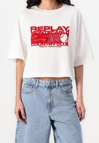 Bílý zkrácený tričko s červeným grafickým potiskem s nápisem "REPLAY MONTECABLO 250 COUNTRY CLUB." Má kulatý výstřih a široké rukávy. Viditelné jeansové šortky.