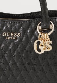 Borsa a tracolla nera con motivo trapuntato realizzata in materiale lucido. Presenta un logo dorato "GUESS" e un ciondolo con le lettere "GS". Dettagli includono una chiusura con zip e manici robusti.