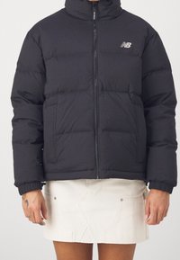 Osoba ubrana w czarną kurtkę puchową z zamkiem z przodu i kieszeniami, zestawioną z białą mini spódnicą dżinsową.