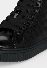 Gros plan d'une basket noire brillante avec motif matelassé sur le côté, semelle thick texturée et lacets noirs passés dans des œillets métalliques.