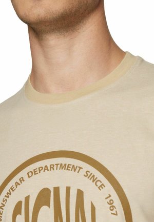 Close-up af en person iført en beige t-shirt med rund halsudskæring og et brunt cirkulært logo med teksten "MENSWEAR DEPARTMENT SINCE 1967."