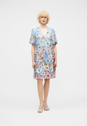 Kvinde med krøllet blondt hår iført en knælængde, lyseblå kjole med flerfarvede blomsterprint og puffede korte ærmer, stående oprejst.