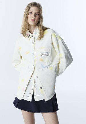 Junge Frau mit langen blonden Haaren trägt eine weiße übergroße Jacke mit bunten Details, einem Aufnäher mit der Aufschrift „MERCI“ und einem schwarzen Rock.