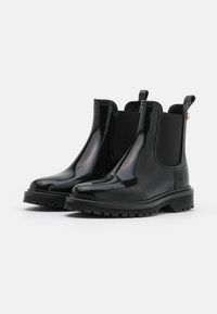 Botas Chelsea de goma negra y brillante con paneles laterales elásticos, suela texturizada negra y una lengüeta en la parte trasera para facilitar su colocación.