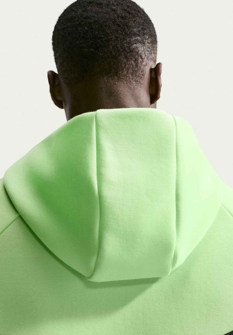 Mand med kort sort hår iført en lysegrøn hættetrøje, set bagfra mod en ensfarvet hvid baggrund.