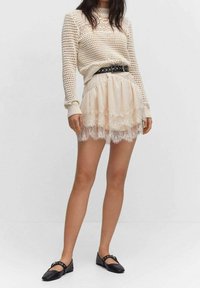 Suéter de punto crema con un patrón texturizado, combinado con una falda beige en capas con borde de encaje y zapatos planos negros con detalles de hebilla.