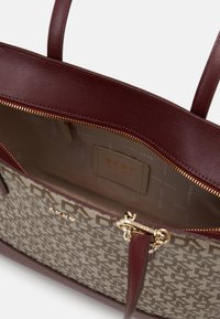 Sac cabas DKNY marron et beige avec motif monogramme, garniture en cuir bordeaux, fermeture éclair dorée et étiquette intérieure indiquant "DKNY New York".