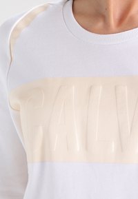 Sudadera blanca con una banda texturizada en crema y letras en relieve que dicen "CALV" en el pecho. Tela suave con mangas raglán.