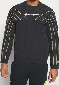 Top sportivo nero con linee gialle a contrasto, caratterizzato da un collo rotondo e una vita elastica. Include il logo Champion sul davanti.