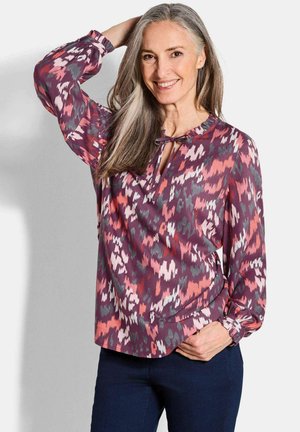 Langemouwen, kleurrijke blouse met een bloemenpatroon in tinten roze, paars en groen; heeft een keyhole-halslijn en gathered manchetten.