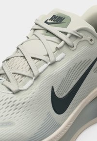 Κοντινό πλάνο ανοιχτόχρωμου γκρι παπουτσιού τρεξίματος Nike με υφή υφάσματος, μαύρο λογότυπο Swoosh και λευκά κορδόνια δεμένα πάνω σε μαξιλαρωμένη γλώσσα με την επιγραφή "RUN".