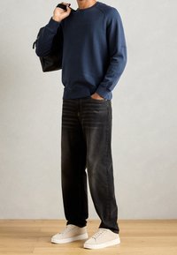 Uomo che indossa un sweatshirt blu navy, jeans neri, sneakers bianche, con uno zaino nero a tracolla, in piedi su un pavimento in legno.