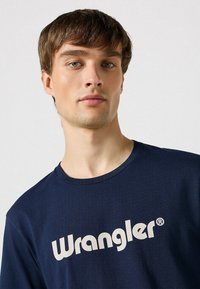 Mørkeblå bomulds T-shirt med rund hals, prydet med et stort hvidt "Wrangler" logo i fed skrift på brystet.