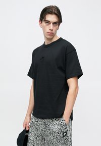 Młody mężczyzna ubrany w czarną koszulkę Nike i czarno-białe wzorzyste szorty, trzymający czarną czapkę w jednej ręce, stojący na tle jednolitego tła.