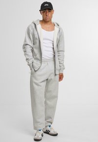 Grauer Kapuzenpullover mit Fronttaschen, kombiniert mit passenden Jogginghosen. Schwarze Kappe mit Logo, weißes Tanktop und Sneakers mit Streifen.