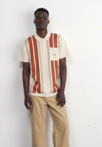 Camisa de manga corta, con cuello, en color crema con rayas verticales en naranja óxido y beige. Presenta un bolsillo en el pecho y un corte relajado. Combinada con pantalones beige claros.