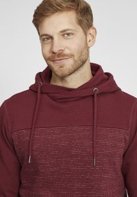 Felpa bordeaux con una parte superiore testurizzata e un motivo a righe orizzontali. Presenta un cappuccio con cordoncino e dettagli metallici.