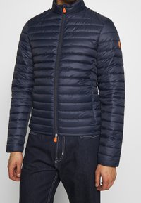 Marineblå dunjakke med vandret quiltning, høj krave, lynlås i fuld længde med orange trækflig og en rund logo på ærmet.